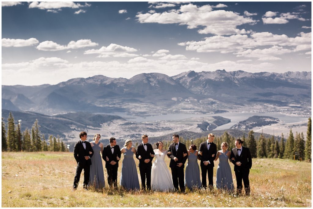 bridal party wearing black tuxes and soft blue dresses pose in front of mountains in timber ridge lodge in keystone colorado