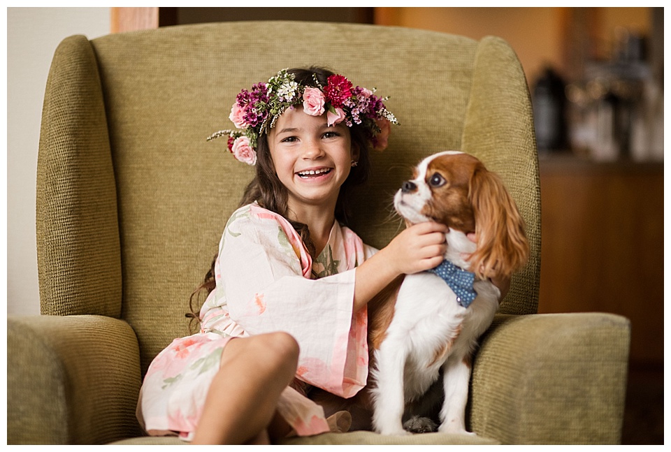 flower girl wearing pink silk robe and flower crown petting a king charles spaniel dog 