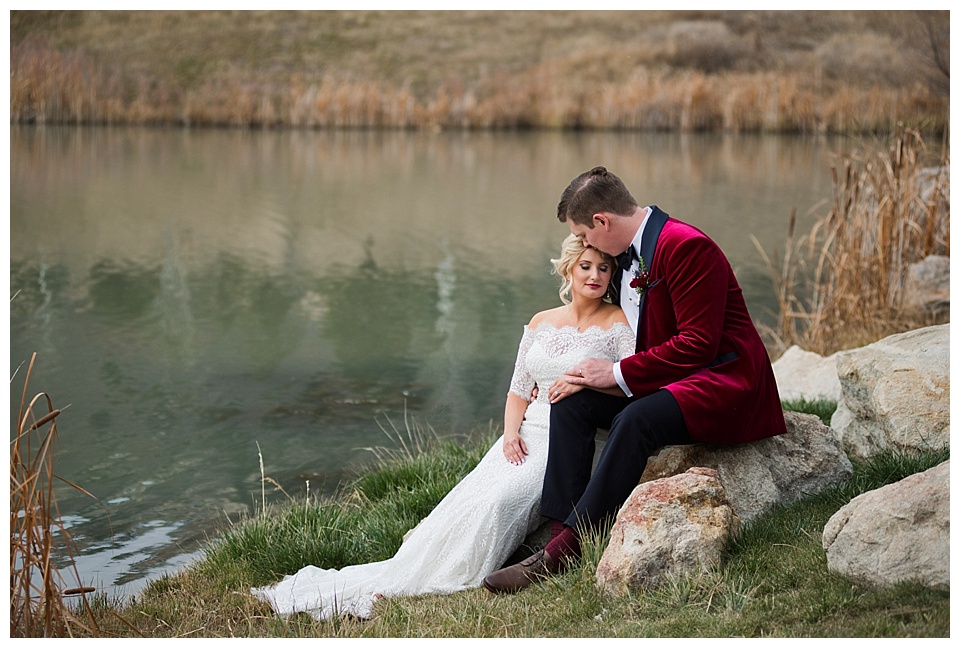 bride and groom sit on rock by lake at spruce mountain ranch with red velvet jacket