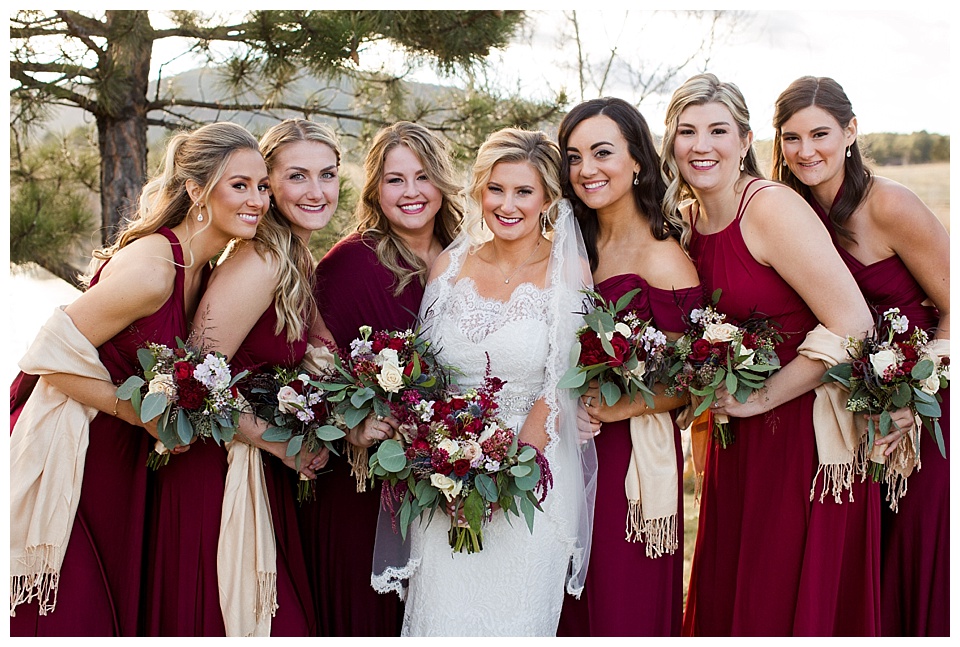 bride poses with bridesmaids wearing red dresses and tan shawls 