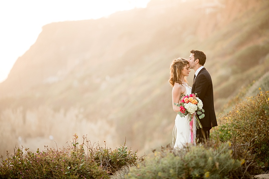 groom kisses bride's head in front of stunning sea cliff backdrop