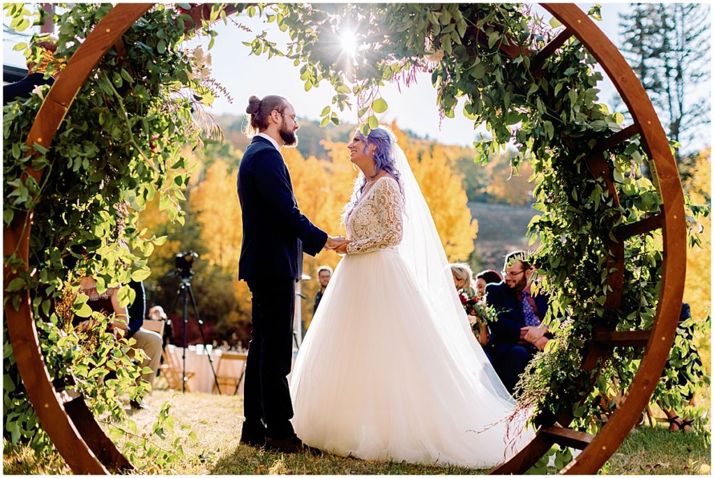 Bride and groom under floral arch designed by Bloom Flower Shop for wedding ceremony at Anderson's Cabin in Beaver Creek.