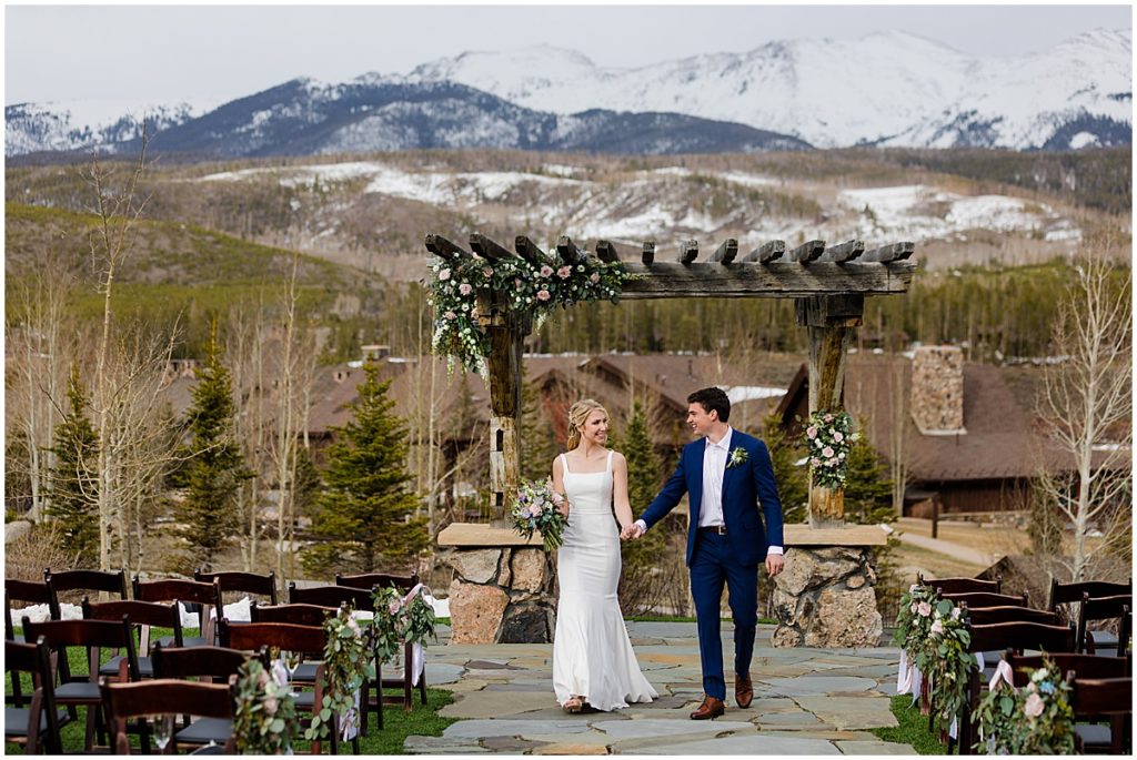 Bride and groom holding hands under alter with snowy mountain tops after wedding ceremony at Devil's Thumb Ranch