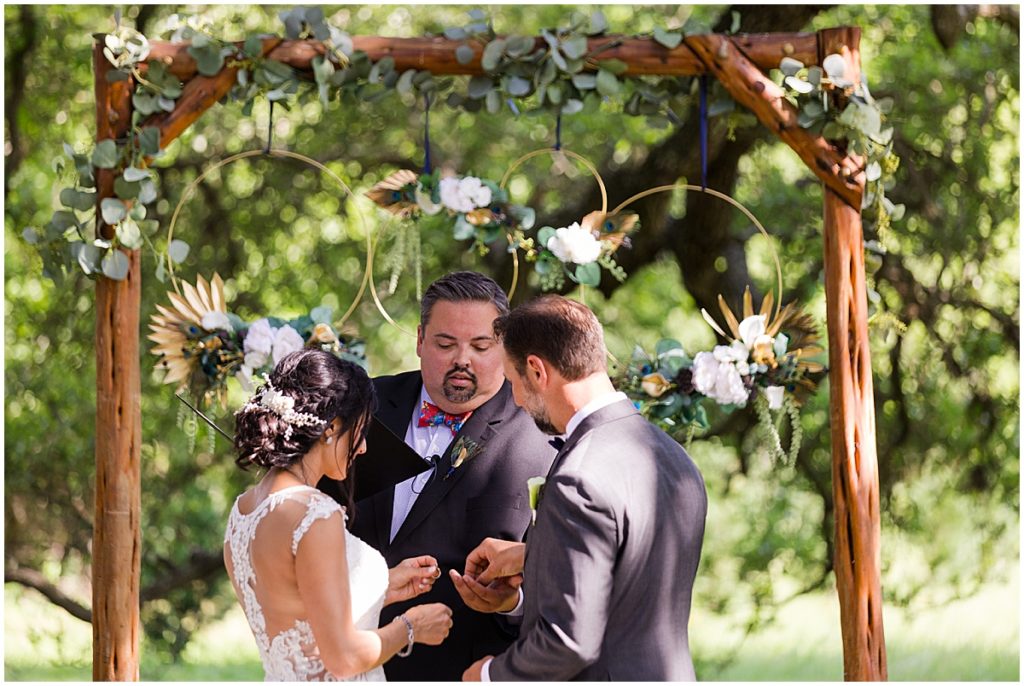 Bride and groom during ceremony outside at The Vineyards at Chappel Lodge standing under arch with floral decor designed by Exquisite Petals.
