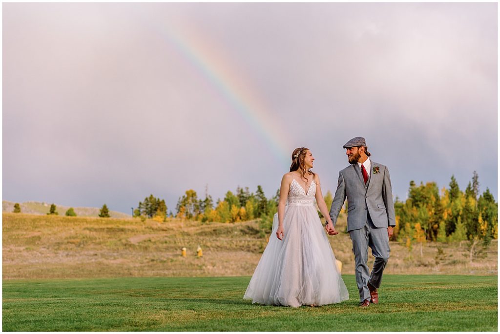Bride and groom on grass field under a rainbow after wedding ceremony at Frisco Day Lodge  Bride is wearing dress from A&BE Bridal Shop and groom wearing suit from Jim's Formal Wear. 